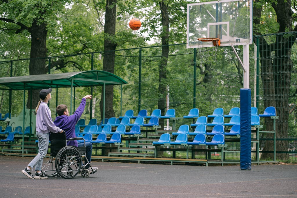A person in a wheelchair playing basketball with a friend outdoors on a court.